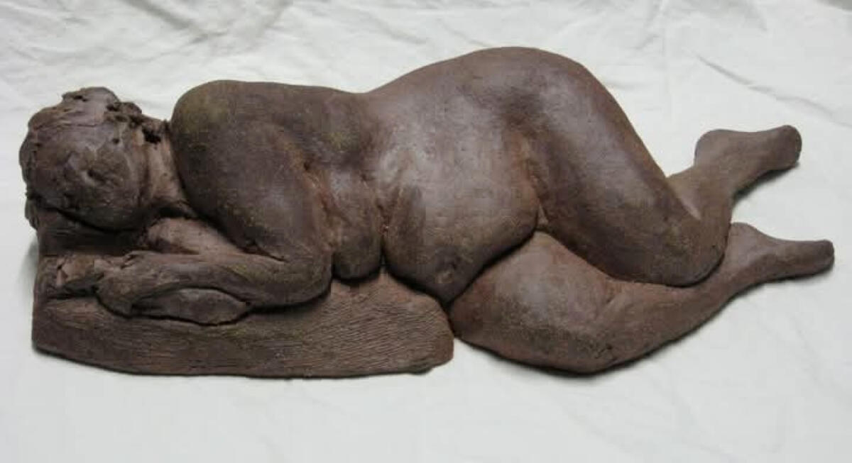 Reclining figure Reclining figure of a nude woman laying on her right side by Shane Brodie. She has a full belly and thighs and wide hips. Facial features are less distinct or detailed. The surface of the piece is a dark matte brown color.