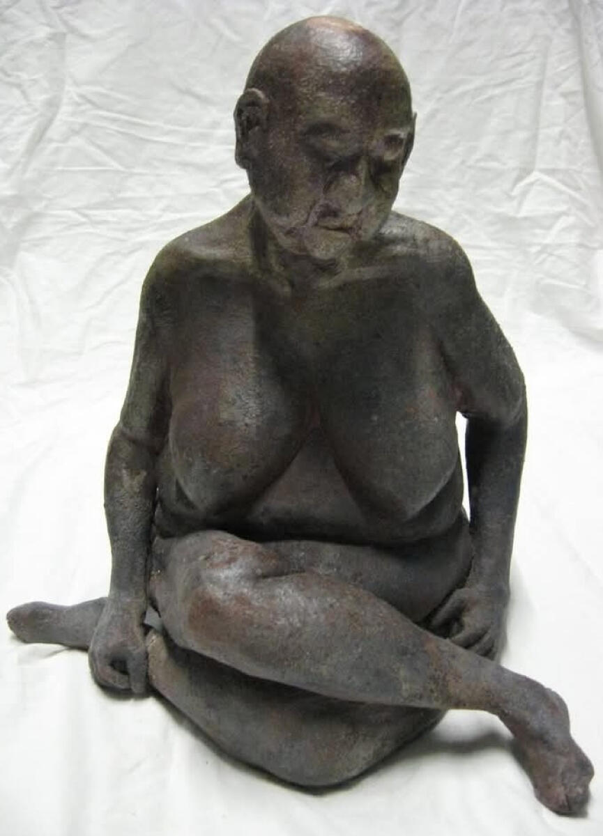 Angle of Repose Woofired sculpture of an seated older female figure by Shane Brodie. The surface is a mottled dark grey with a blush of red from a heavy reduction environment. The figure is sitting upright with legs crossed and arms down at the sides.